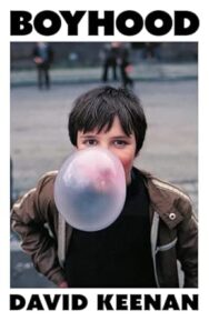 Image of a book cover featuring a photograph of a young brown haired boy outdoors, centred against a blurred urban background. The boy is blowing a large, nearly translucent pink bubble of chewing gum that obscures much of their face. He wears a brown jacket with a zip. The image is framed by a thick white border. The title “BOYHOOD” appears in bold black uppercase letters at the top of the border, and the author’s name “DAVID KEENAN” appears in matching black text at the bottom.